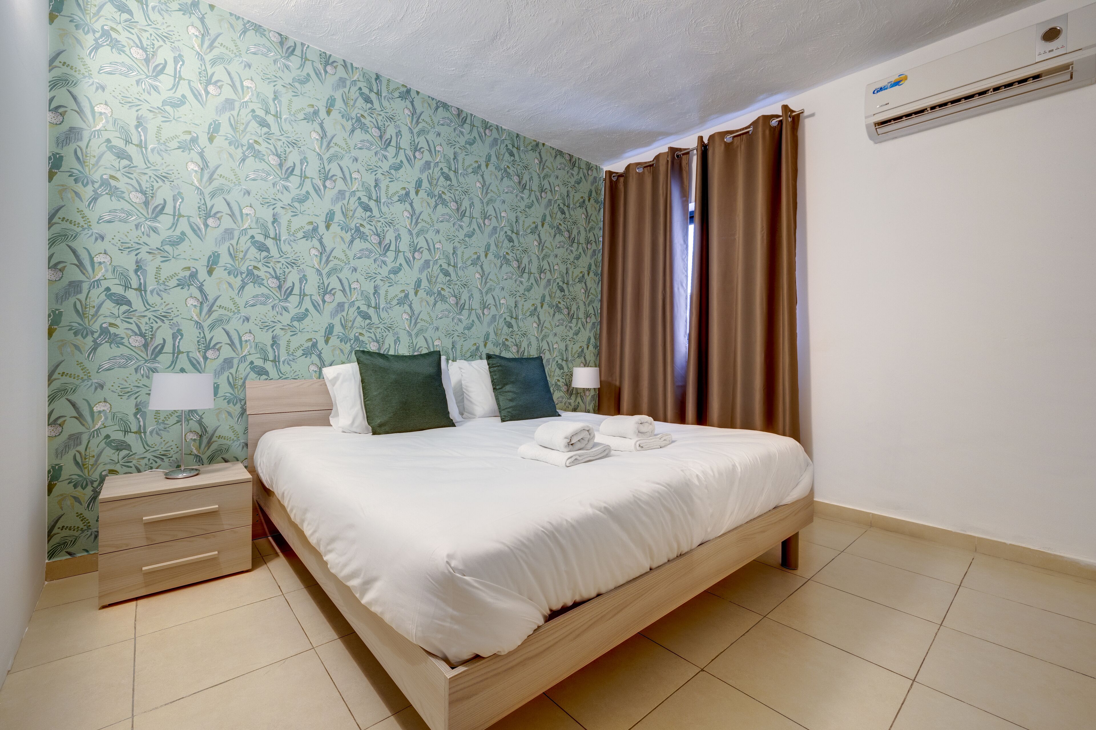A neatly made bed with white linens, a wooden nightstand with a lamp, and a floral-patterned wall.