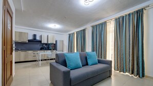 A modern living room with a grey sofa, two blue cushions, a dining table with chairs, and a kitchen area in the background. - Sliema Tigne Suites (Sliema)
