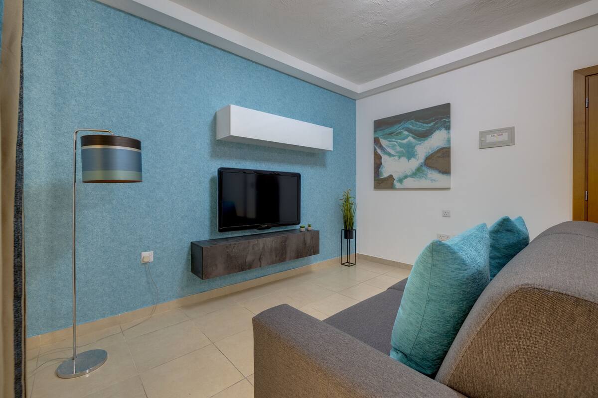 A modern living room with a flat-screen TV mounted on a wall, a floor lamp, and a sofa with cushions.