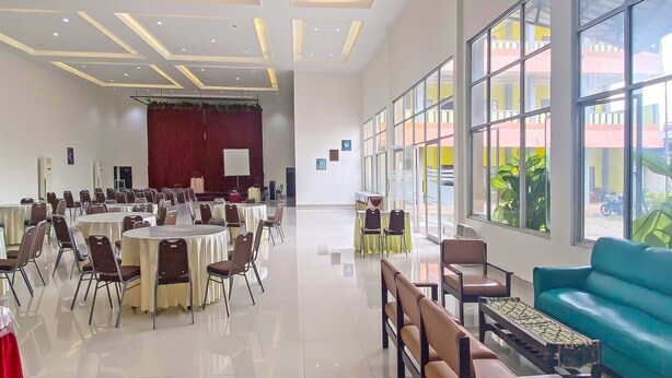Una amplia sala de conferencias con mesas y sillas redondas, un escenario con cortina roja y grandes ventanales que ofrecen vistas al exterior.