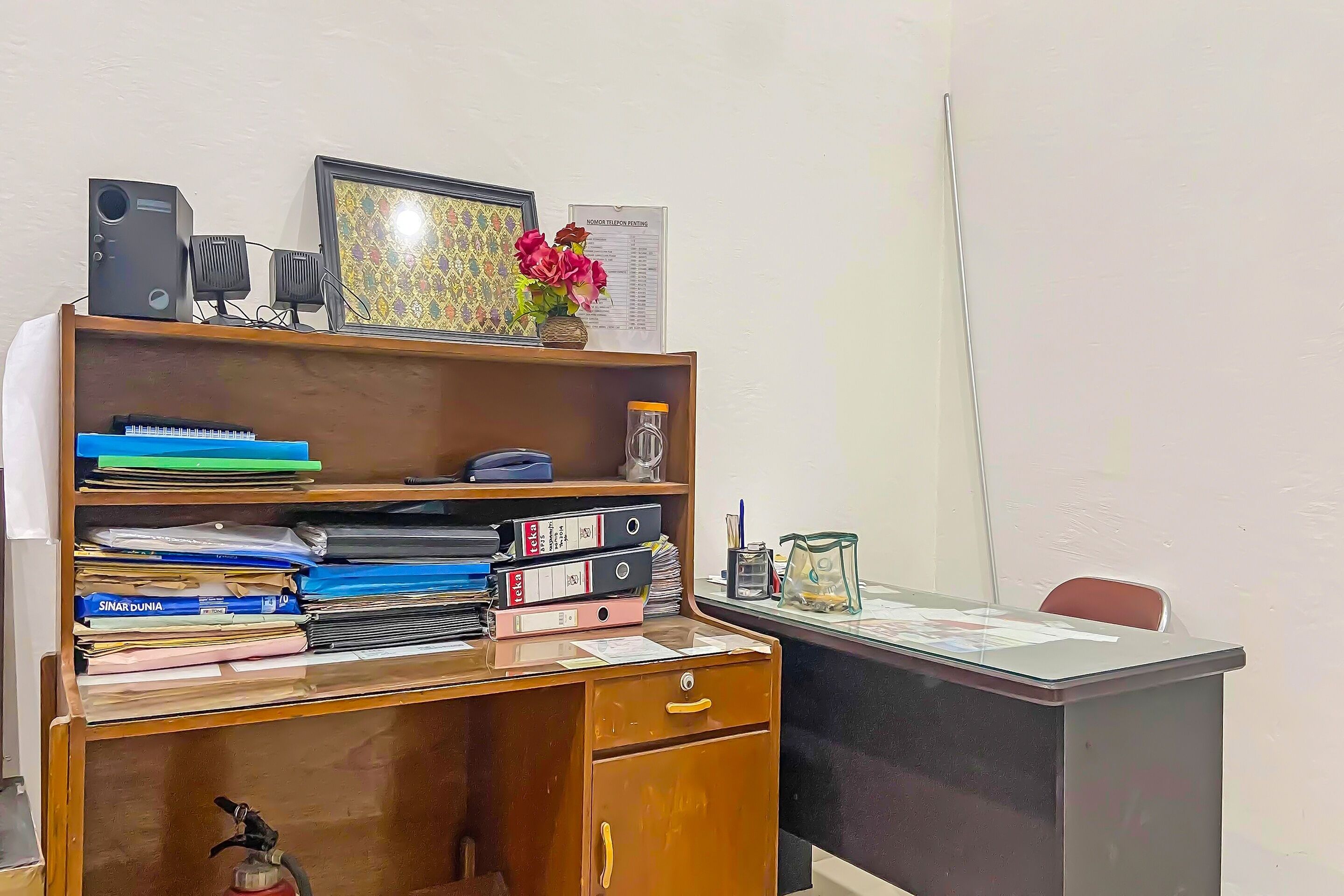 A wooden desk with a computer, a monitor, and a vase with flowers. There are shelves with binders and a framed picture.