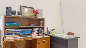 A wooden desk with a computer, a monitor, and a vase with flowers. There are shelves with binders and a framed picture.