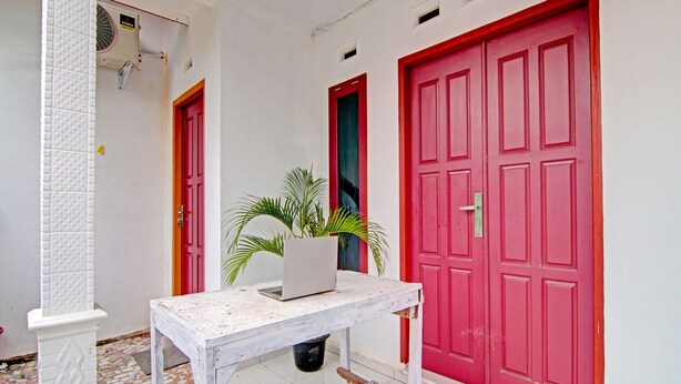 A hallway with a white tiled floor, a white table with a laptop and a potted plant, and two red doors. - SPOT ON 91950 Guest House TekNong Syariah (Bangkinang)