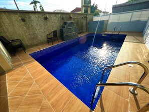 A swimming pool with a tiled deck, a metal ladder, and a wall with potted plants.