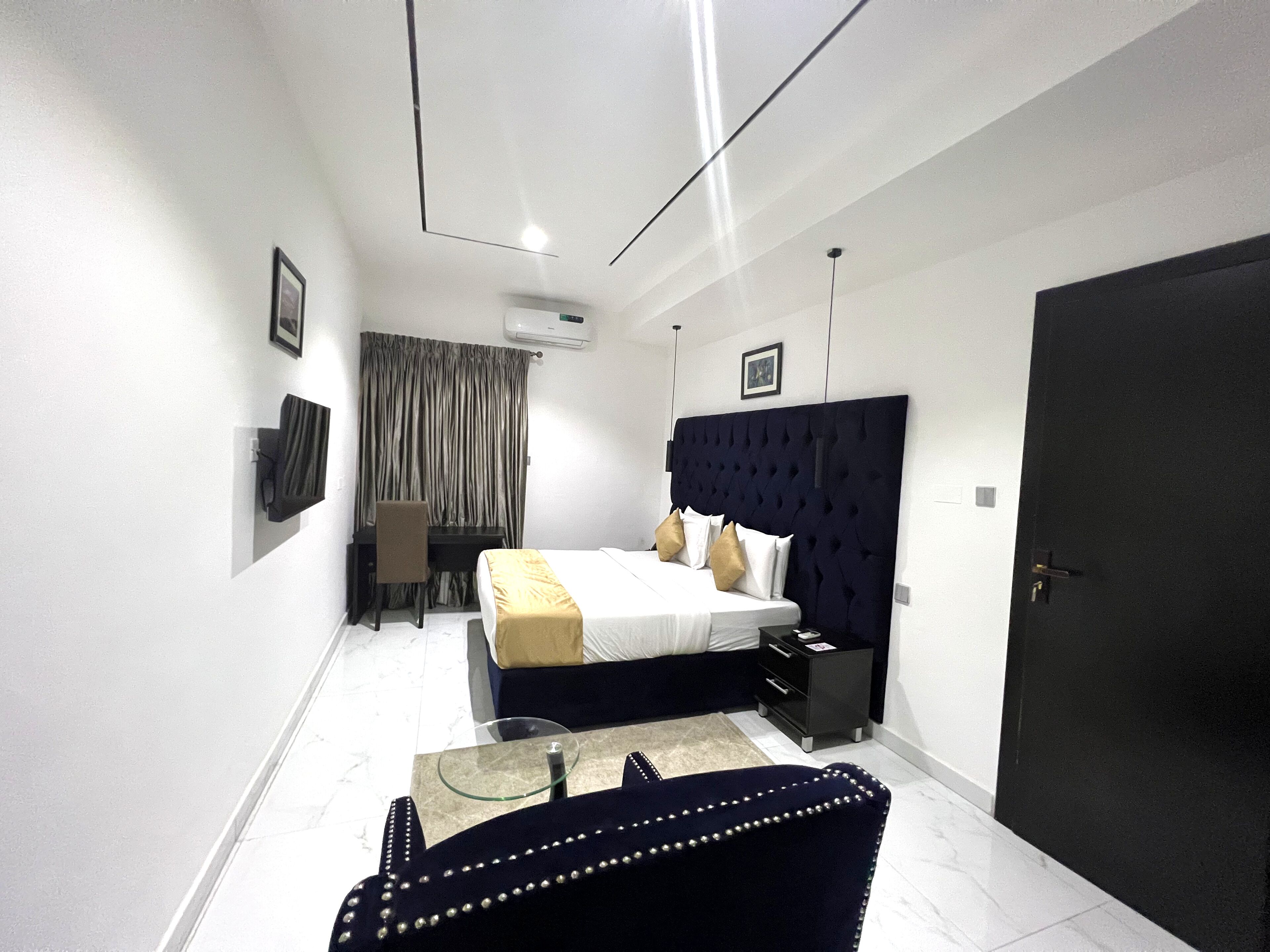 Quarto de hotel moderno com uma cama, uma escrivaninha, uma cadeira, uma mesa de centro de vidro e um sofá de cor escura.