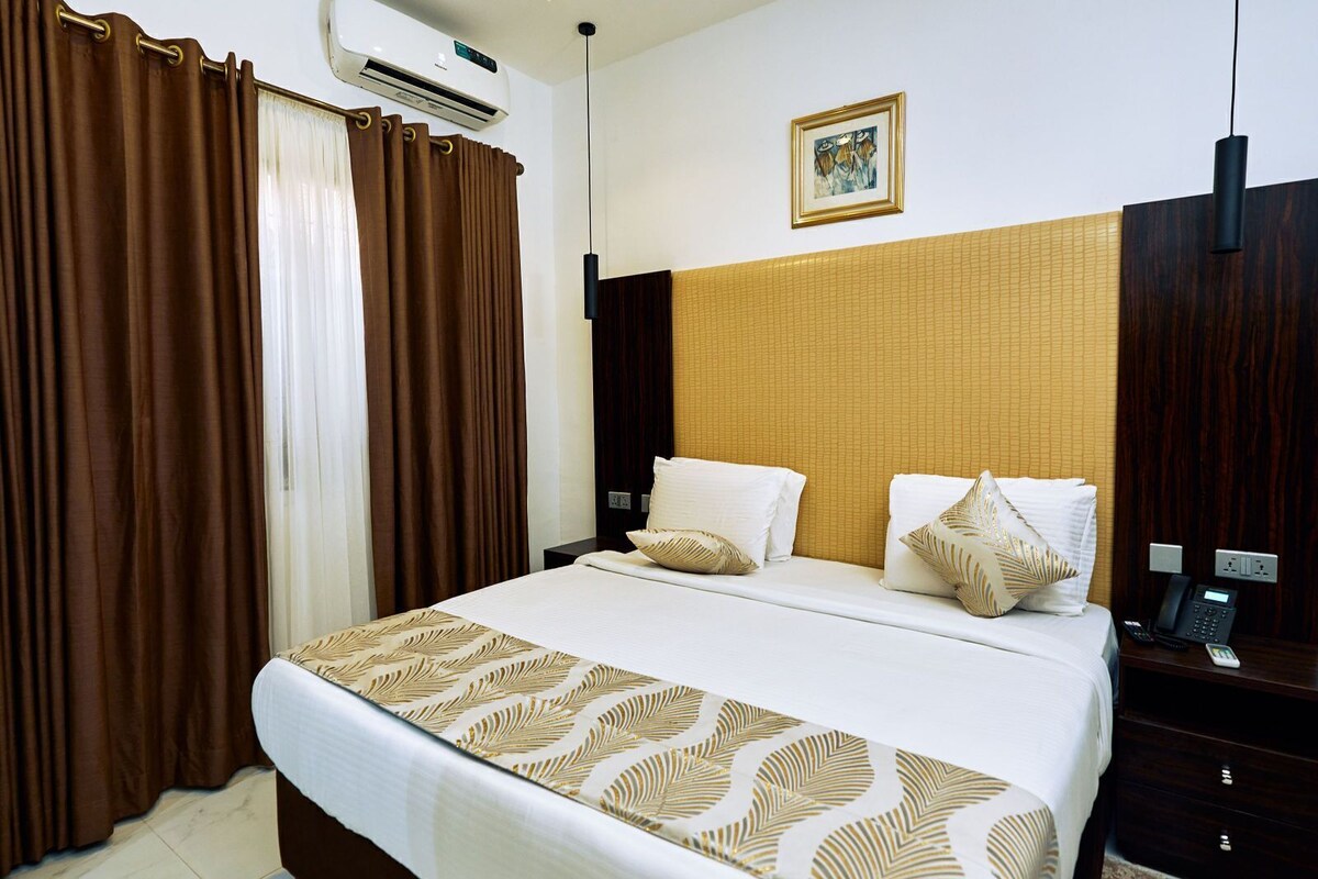 A hotel room with a bed, brown curtains, a wall-mounted air conditioner, a painting, and a bedside telephone.