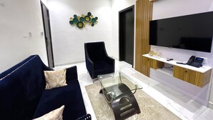 A modern living room with a dark blue sofa, a glass coffee table, a black armchair, a wall-mounted TV, and a decorative wall piece. - Riviera Lekki Resort (Lekki)