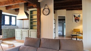 Een woonkamer met een beige bank, houten balken in het plafond, een keuken met groene kasten en een wandklok.
