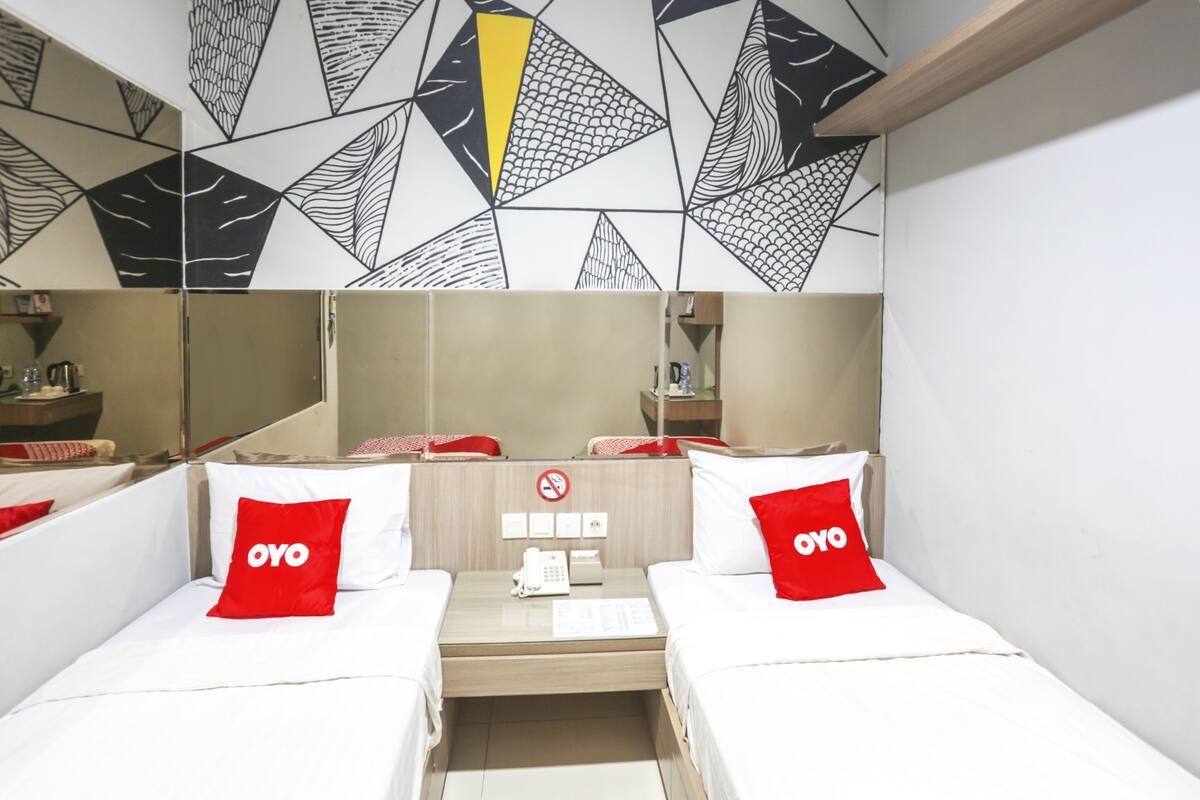 Habitación de hotel con dos camas, cada una con un almohadón rojo con la etiqueta "OYO", una mesita de noche con un teléfono y una lámpara, y una pared con un diseño geométrico sobre las camas.