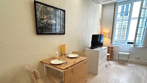 A small, well-lit room with a dining table set for two, a television, and a window with a view of buildings.