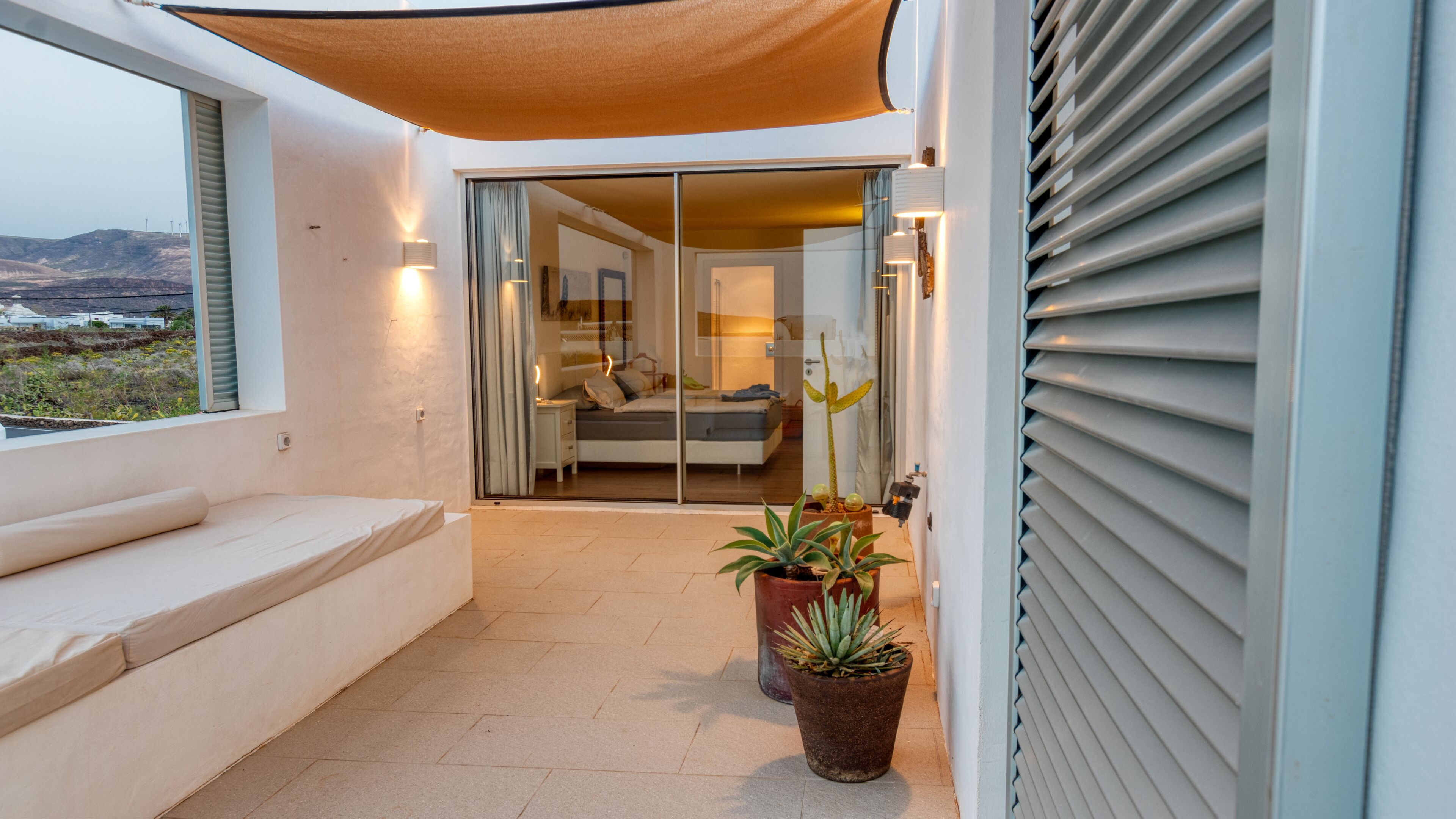 Ein modernes Schlafzimmer mit einem großen Bett, einer Couch und einer Glasschiebetür, die auf einen Balkon führt. Auf dem Balkon stehen zwei Kübelpflanzen.
