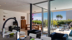 A modern living room with a glass wall, a dining area with a table and chairs, and a kitchen with a sink and a vase of flowers.