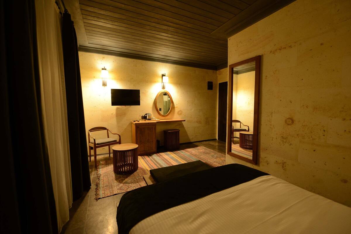 Habitación de hotel con una cama, un escritorio con una silla, una mesita, un espejo y una lámpara fijada a la pared.