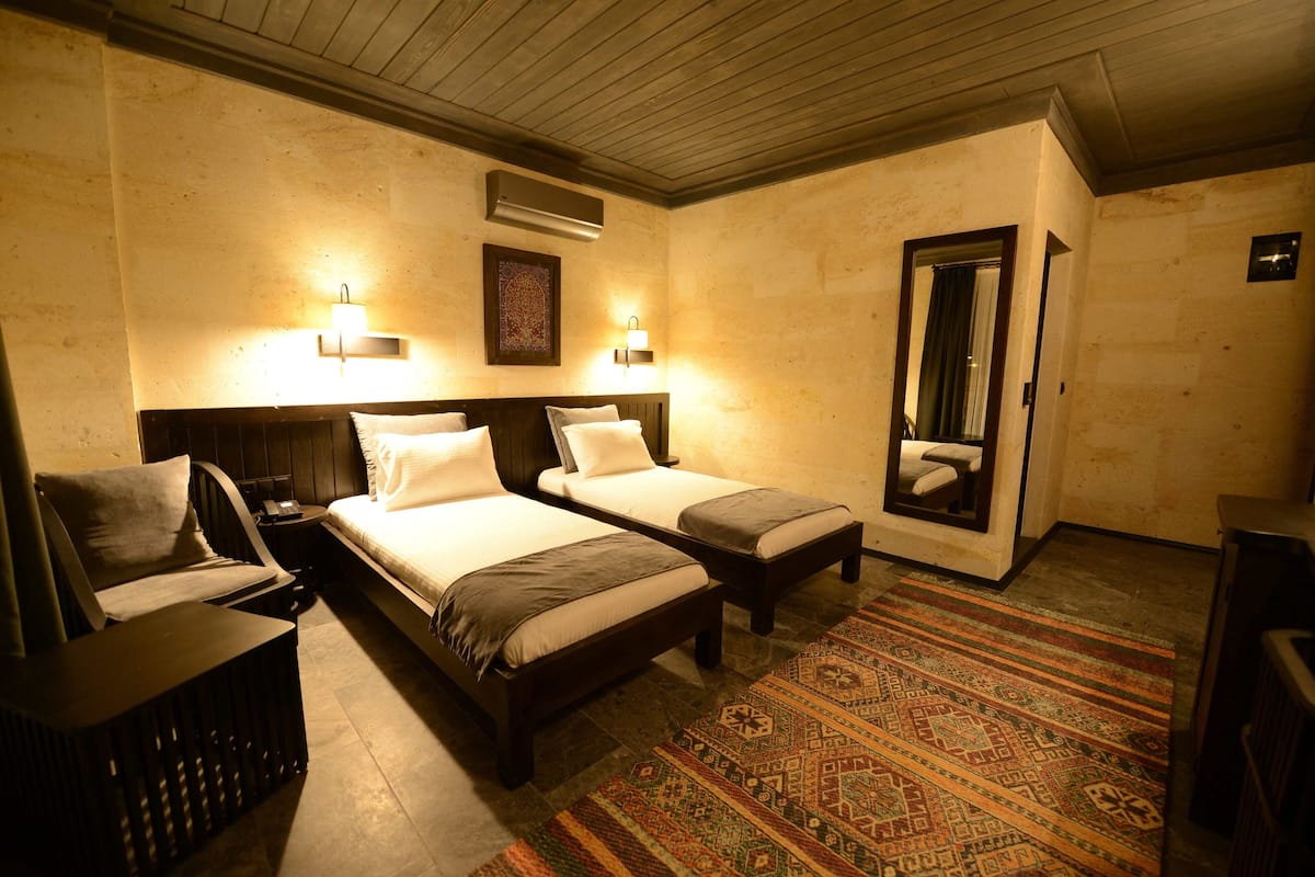 Habitación de hotel con dos camas, una mesita de noche, una pequeña mesa, un banco, un tapete estampado y un espejo.