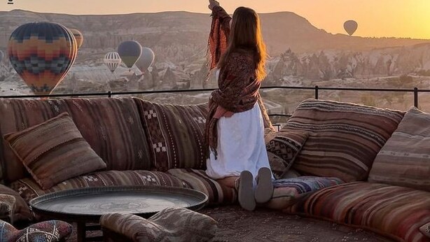 Une personne assise sur un canapé, d’où l’on peut admirer un paysage parsemé de montgolfières.