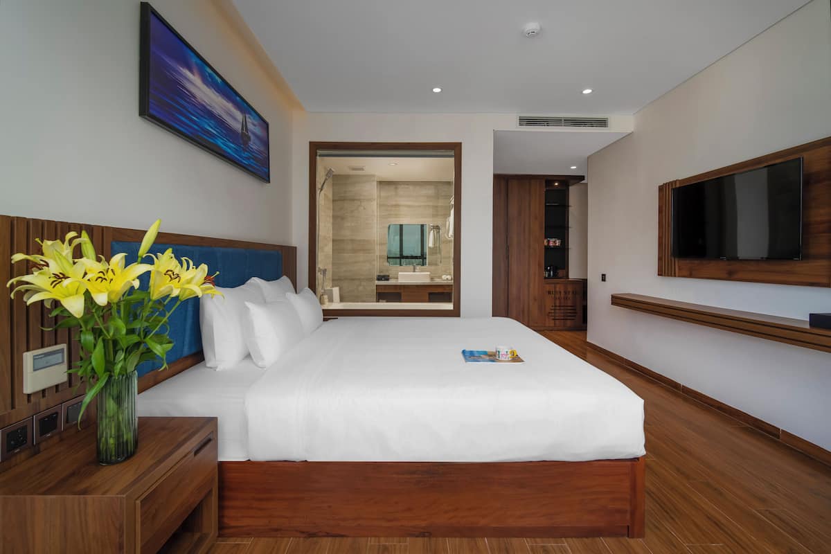 A modern hotel room with a large bed, wooden headboard, a bedside table with a vase of flowers, a TV mounted on the wall, and a bathroom visible through an open door.