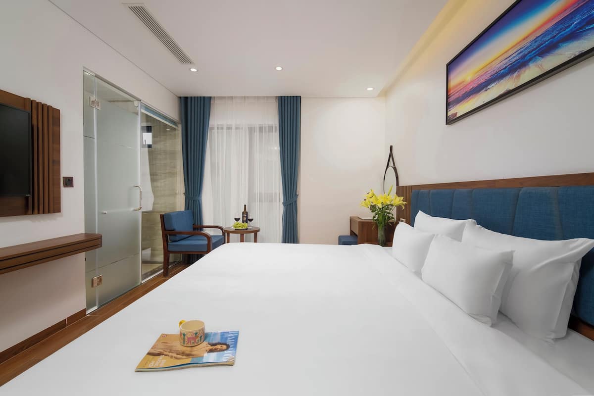 A hotel room with a large bed, a flat-screen TV, a desk with a chair, a small table with a vase of flowers, and a painting on the wall.
