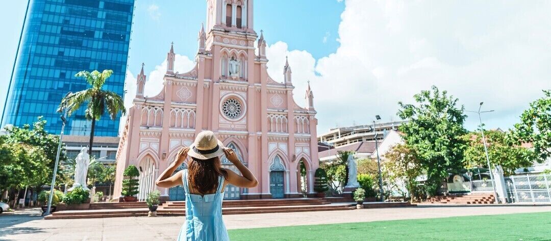 A person in a light blue dress and hat standing in front of a pink church with a clock tower.