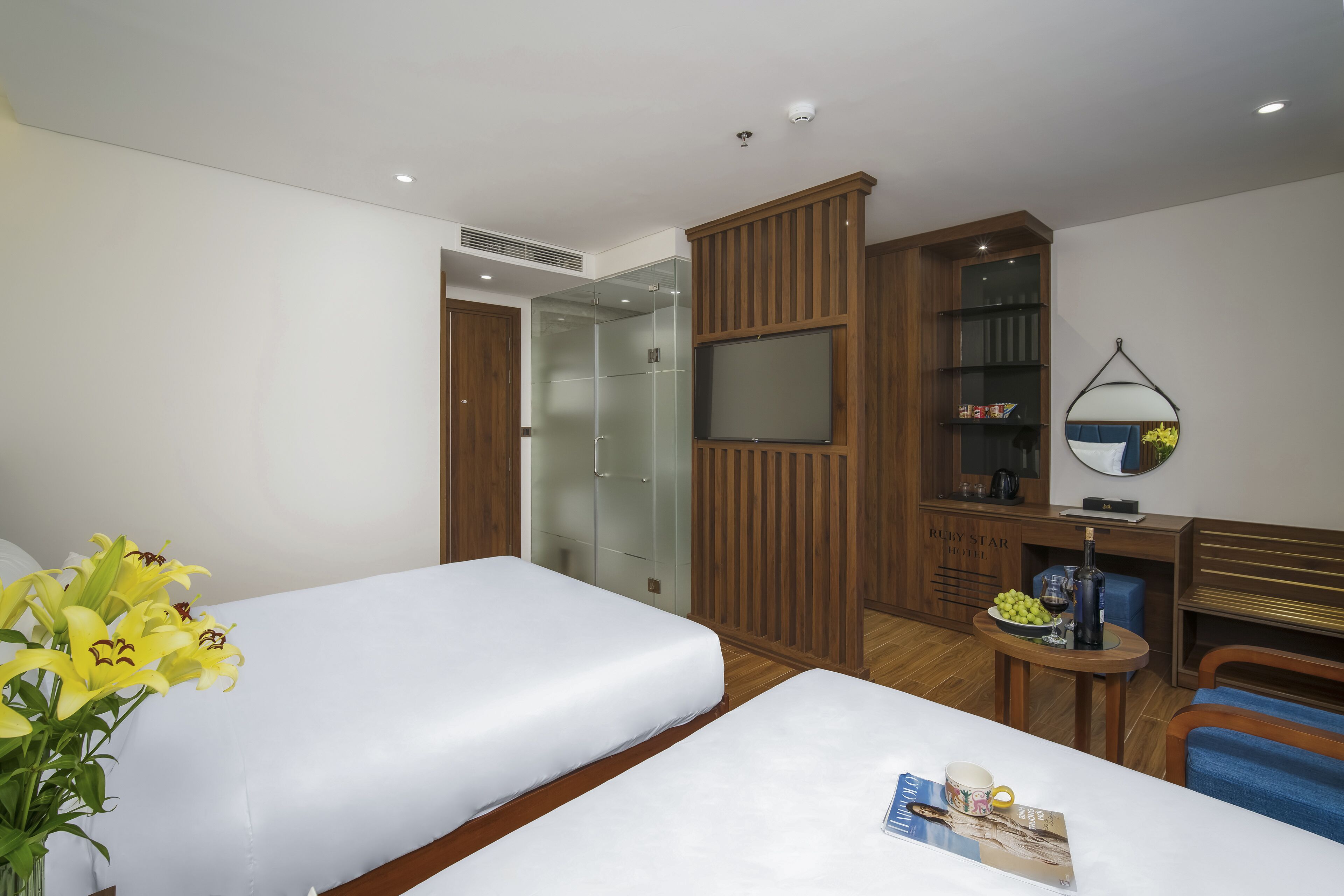 Une chambre d’hôtel avec deux lits, une armoire en bois, une cabine de douche en verre, une petite table avec une bouteille et des verres, et un miroir au mur.