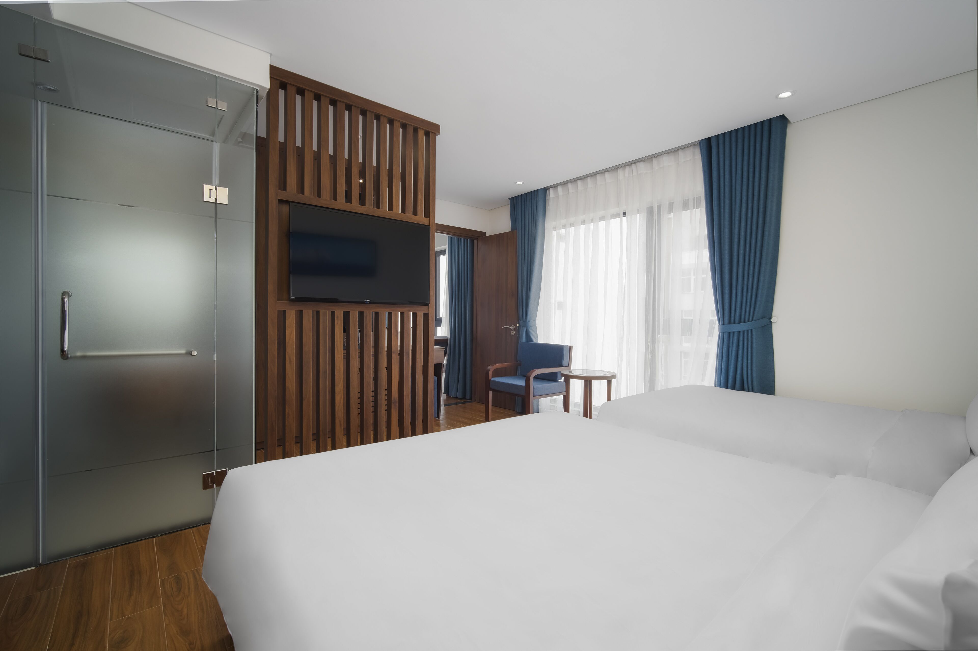 A hotel room with a large bed, a TV mounted on a wooden panel, a desk with a chair, and a window with blue curtains.