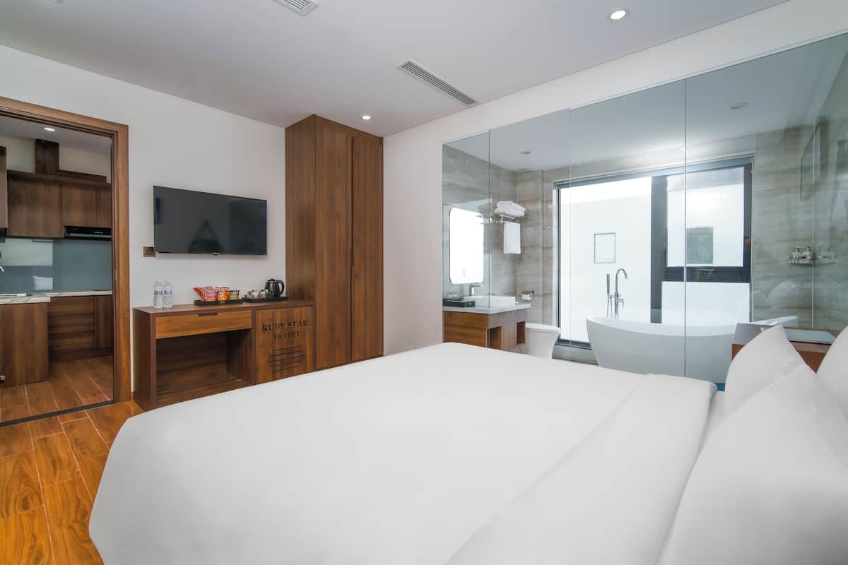A modern hotel room with a large bed, a wooden desk with a flat-screen TV, a glass-enclosed bathroom, and a view of the city.
