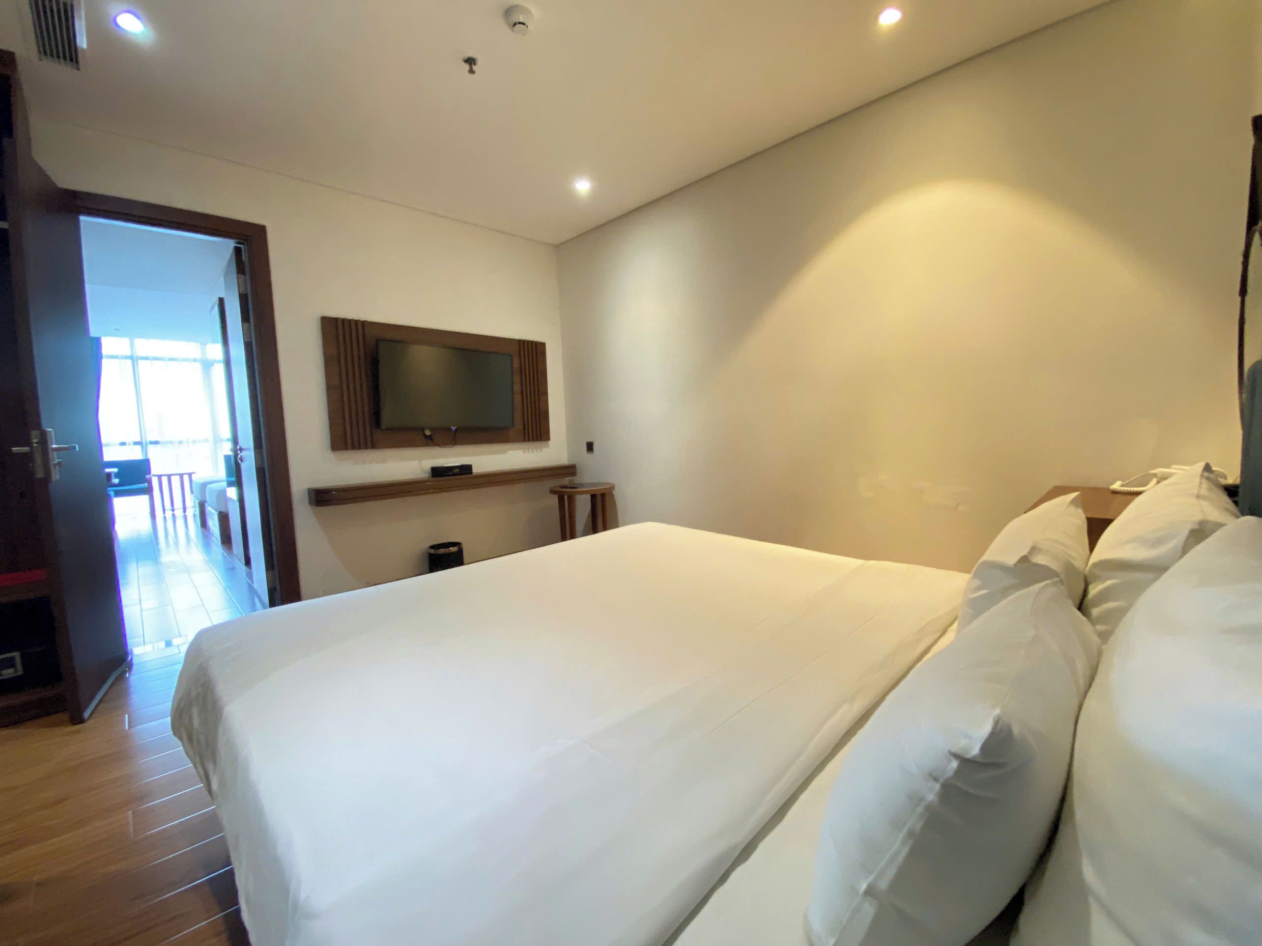 Habitación de hotel con una cama grande, un televisor montado, una mesita pequeña y una puerta que da a un balcón.