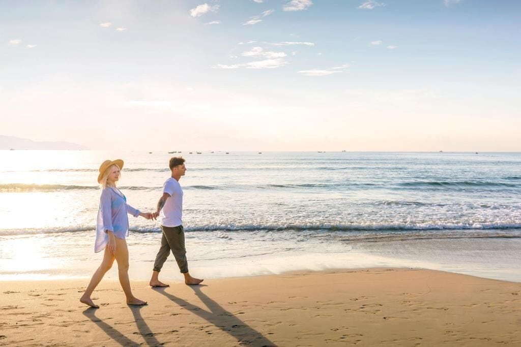 Ein Paar geht Hand in Hand am Strand, im Hintergrund das Meer.