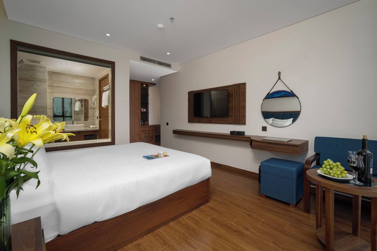 A modern hotel room with a large bed, a bedside table with a vase of flowers, a wall-mounted TV, a mirror, a small table with a bottle and glasses, and a blue chair.