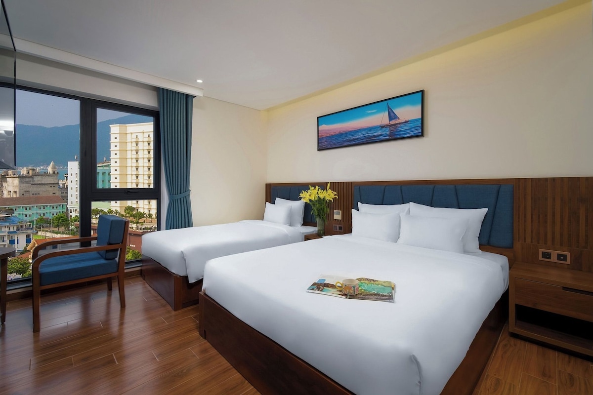 A hotel room with two beds, a large window with a city view, a painting on the wall, and a chair with a view.