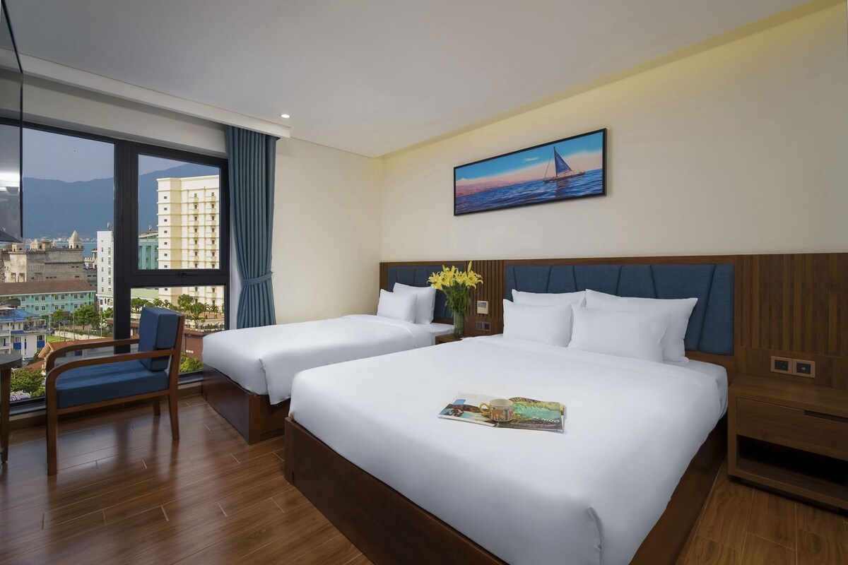 A hotel room with two beds, a large window with a city view, a painting on the wall, and a small table with a magazine.