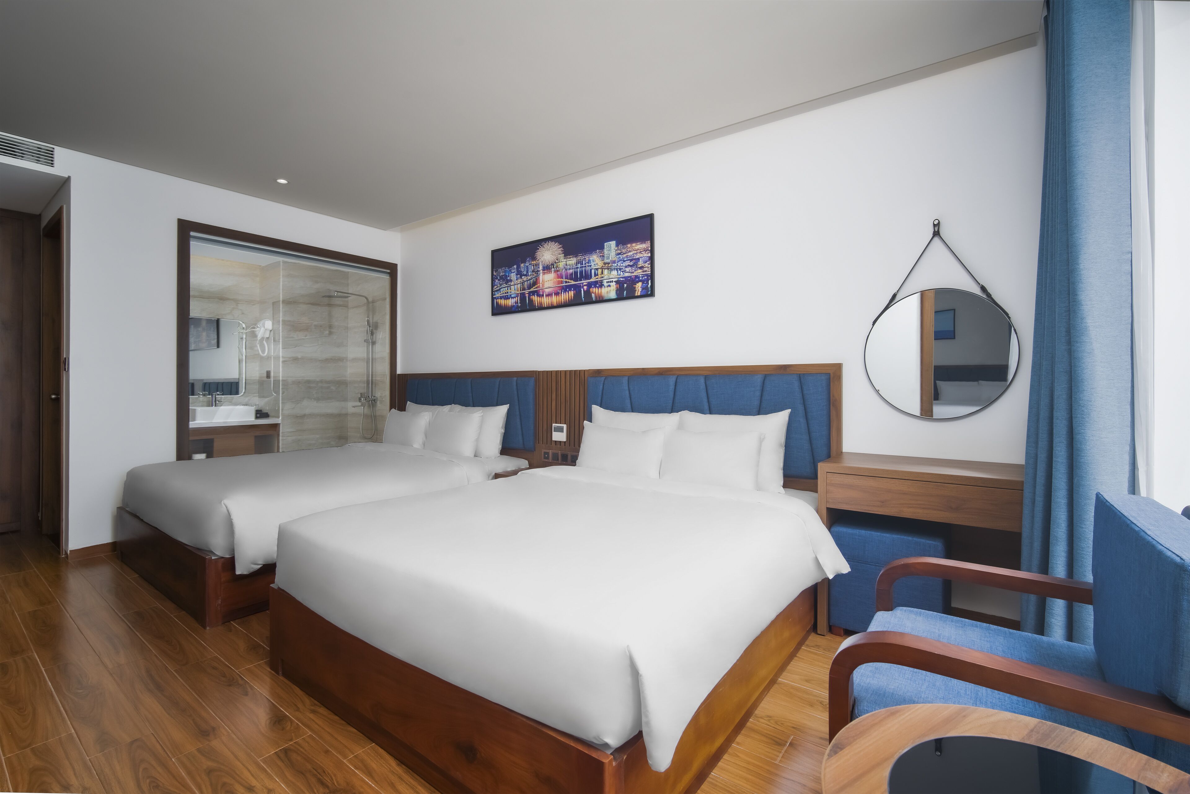 A hotel room with two beds, a wooden floor, a cityscape painting, a round mirror, and a window with blue curtains.