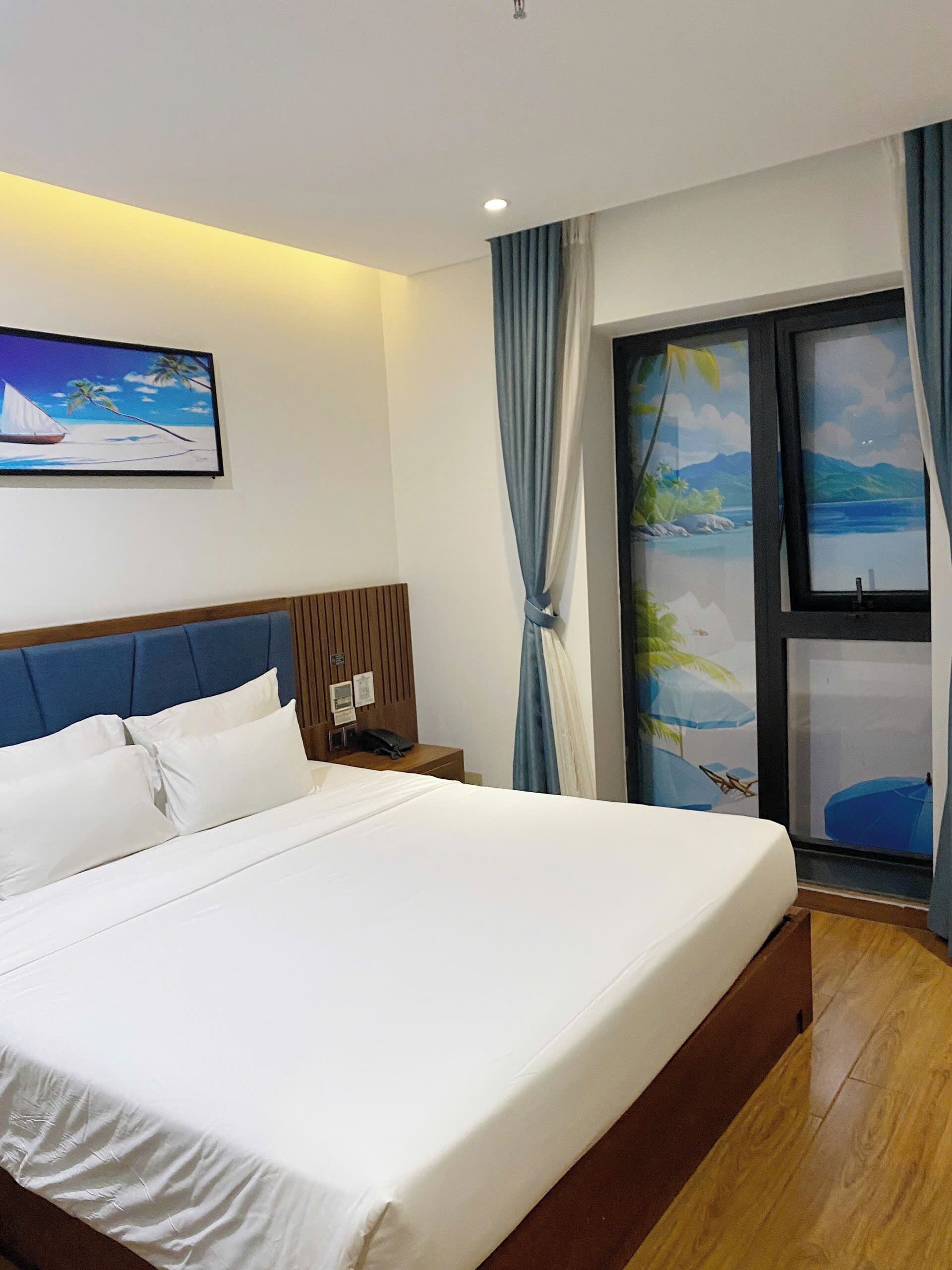 Una habitación de hotel con una cama grande, cabecera de madera, mesita de noche, un cuadro de un velero y vista a una playa a través de una ventana.