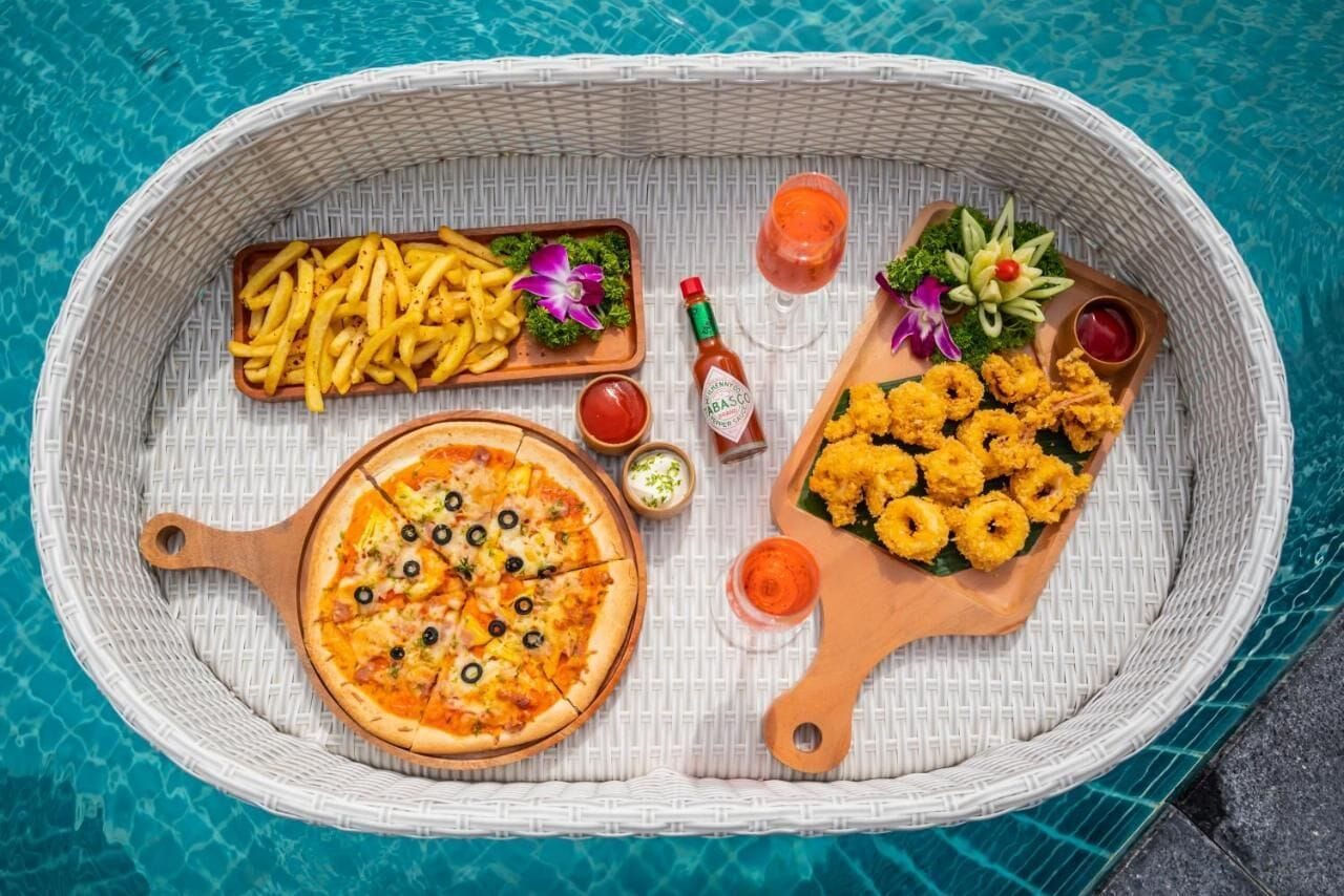A wicker tray with a pizza, fries, and fried calamari, accompanied by condiments and drinks, placed on a poolside setting.