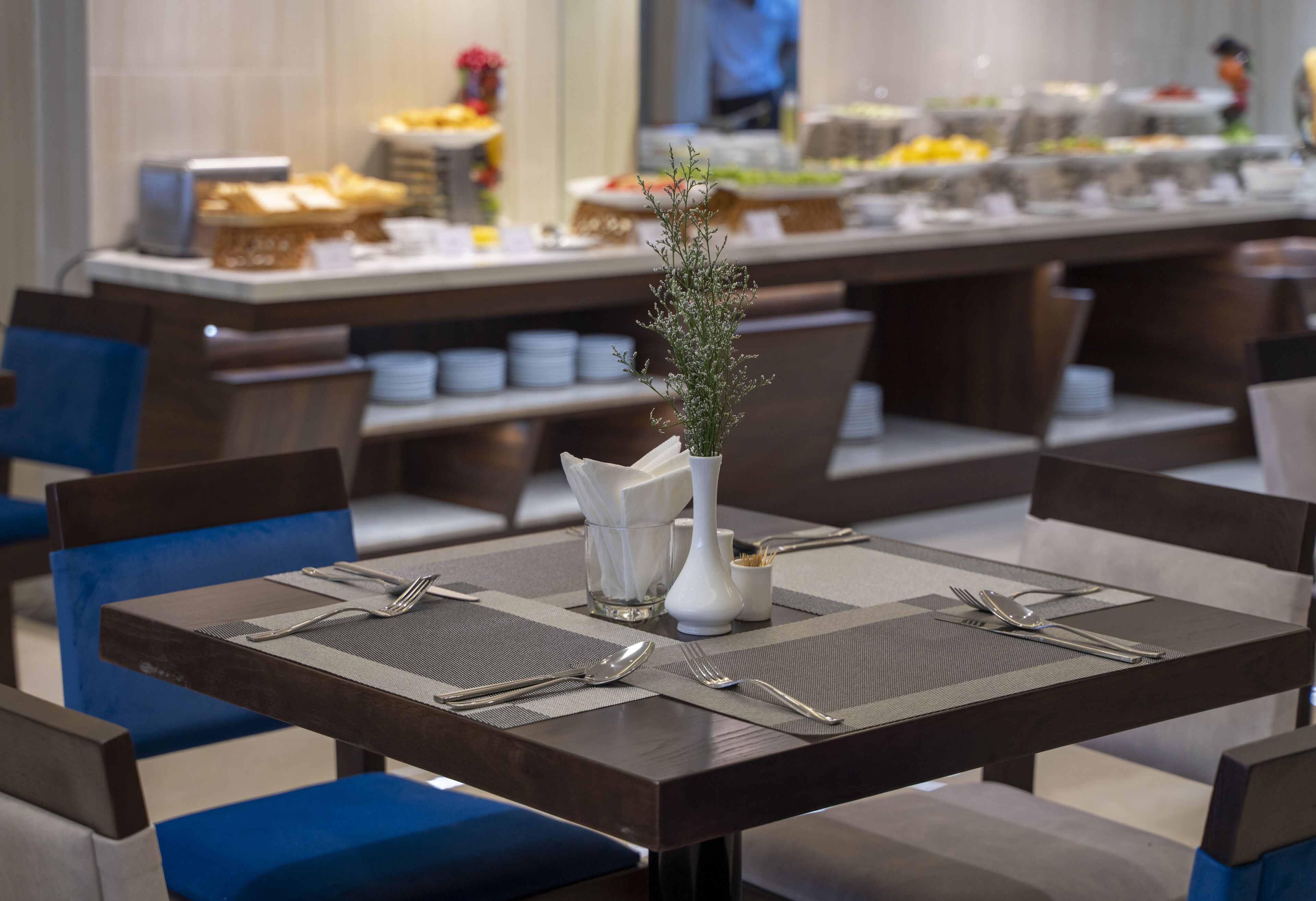 Ein Essbereich mit gedeckten Tischen, Besteck und Servietten, einer Pflanze in einer Vase und einem Buffet im Hintergrund.