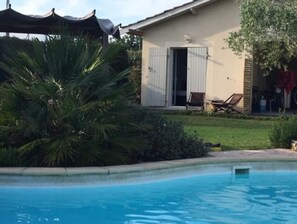 Una piscina con superficie de agua azul cristalina, rodeada por un césped cuidado y un edificio con puerta y ventanas.