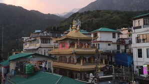 A traditional multi-story building with a golden roof, surrounded by residential structures and mountains in the background. - Hotel Buddha's Abode- Best Hotel in McleodGanj Main Market (Dharamshala)