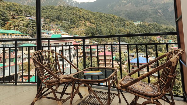 A balcony with wicker chairs and a small table, overlooking a mountainous landscape with buildings in the background. - Hotel Buddha's Abode- Best Hotel in McleodGanj Main Market (Dharamshala)