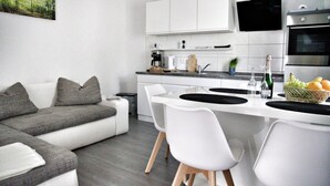 Cuina moderna amb armaris blancs, un televisor negre penjat sobre el forn, i zona de menjador amb cadires blanques i un taulell negre.