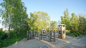 Un parc infantil amb estructures de fusta i un petit edifici de fusta, envoltat d'arbres i un camí pavimentat.