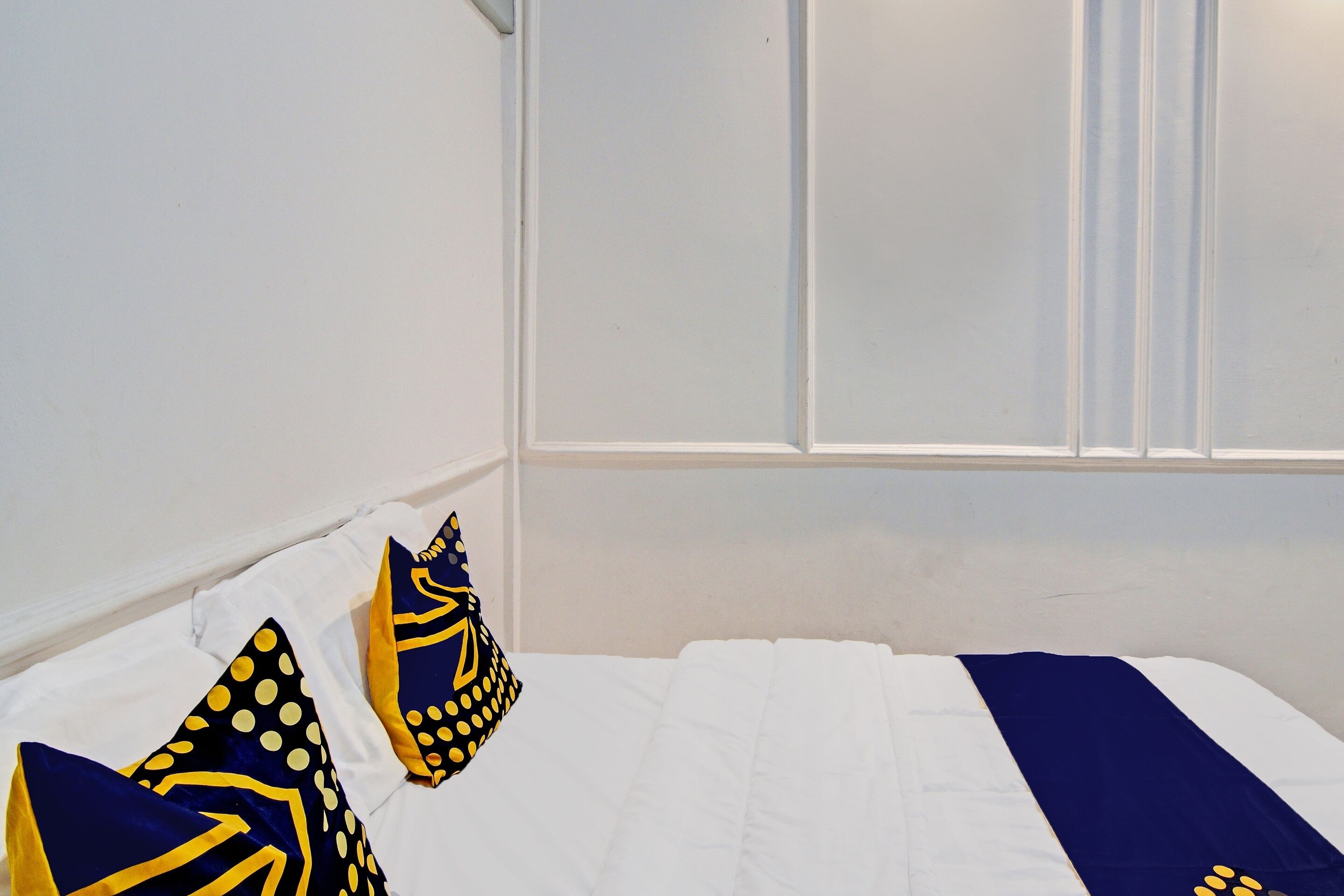 Un dormitorio con cama blanca, almohadas azul marino y amarillas, y una ventana con persianas.