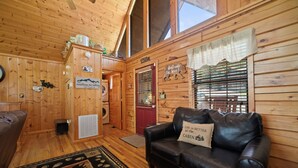 A cabin interior with a black leather couch, wooden walls, a red door, and a window with blinds.