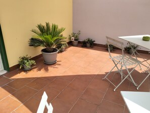 Une terrasse carrelée de carreaux de terre cuite, une table blanche et plusieurs plantes en pot.