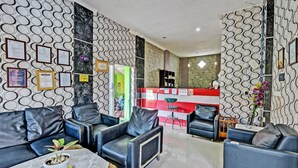 A modern living room with black leather sofas, a coffee table, and a potted plant. There is a reception desk with a red and white striped backdrop in the background. - SPOT ON 91376 Hotel Bintang Flores (Cilacap)