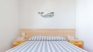 Une chambre à coucher avec une couvre-lit à rayures, une tête de lit en bois, deux tables de chevet avec des lampes et une œuvre d’art représentant une baleine, accrochée au mur.