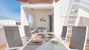 Un espace extérieur lumineux, aménagé pour les repas, avec une table en verre, des chaises blanches et un parasol marron.