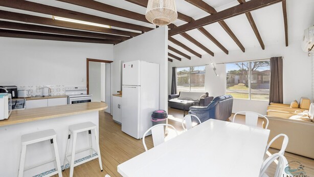 Una cocina moderna con electrodomésticos blancos, un comedor con mesa y sillas blancas, y una sala de estar con sofá y vistas al exterior.