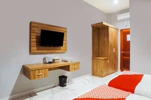 A bedroom with a TV mounted on a wooden panel, a wooden wardrobe, a bedside table with a lamp, and a bed with a patterned blanket.
