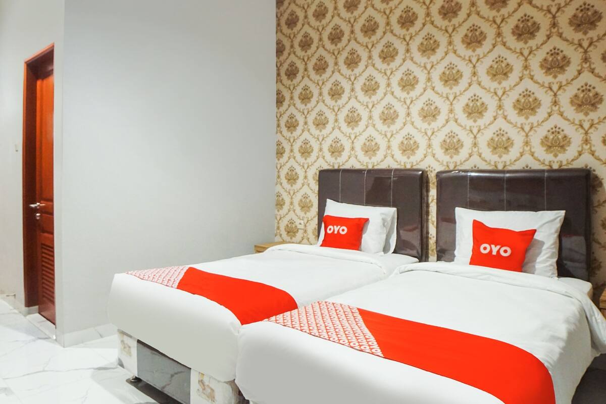 A hotel room with two beds, each with a red and white bedspread and matching pillows. The room has a patterned wallpaper and a wooden door.