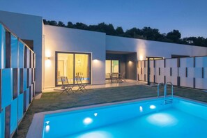 A modern outdoor pool area with a clear blue pool, two lounge chairs, and a glass door entrance to a building. - Deluxe Villa Polydoros (Malevizi)