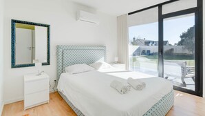 A bedroom with a bed, a bedside table, a mirror, and a window with a view of the outside.